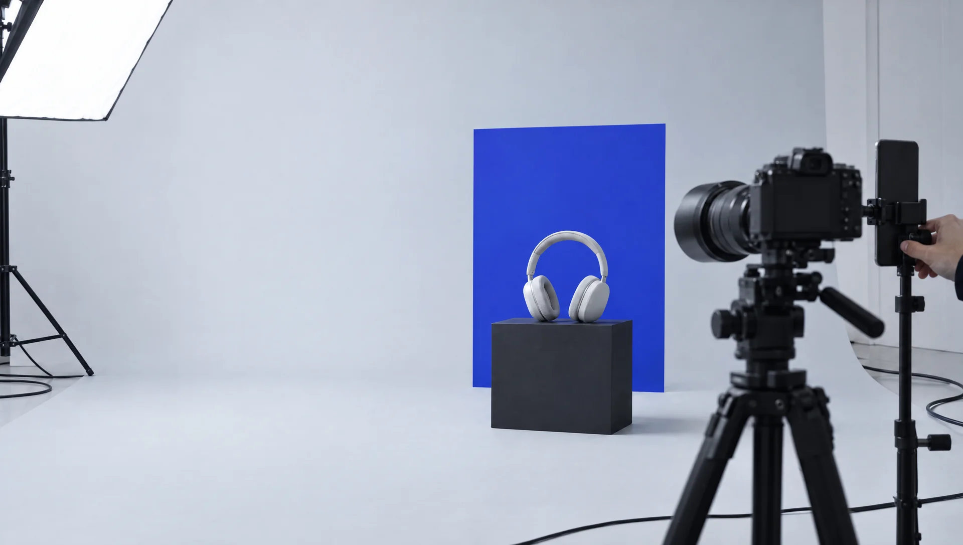A photo studio mid-shoot: pale gray headphones on a black plinth in front of a blue backdrop card.